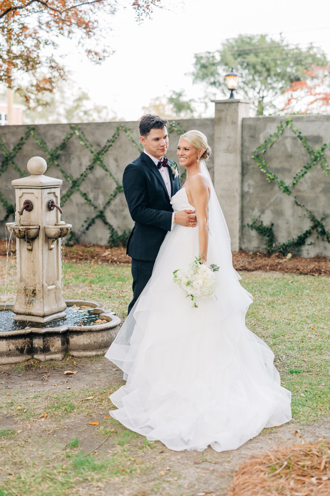 The bride and groom pose in a vertical shot next to a rustic stone fountain, with the bride holding her bouquet at the Hartley House Wedding South Carolina
